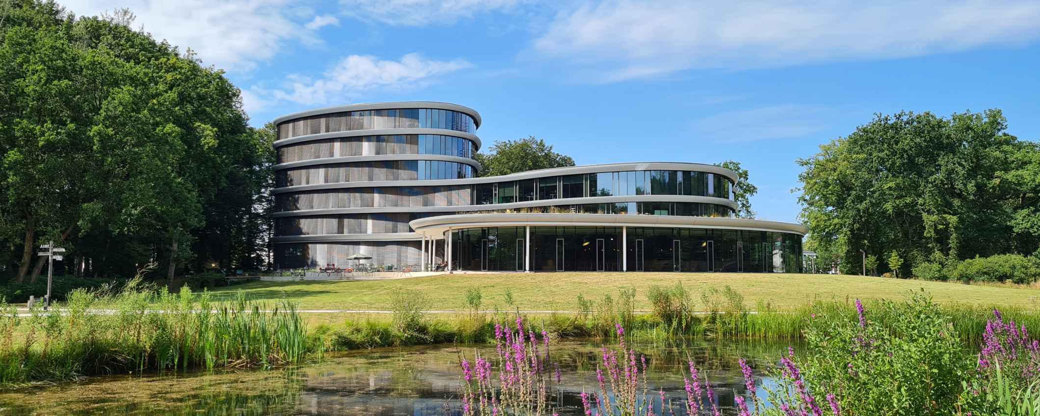 Vooraanzicht van het kantoor van Triodos Bank, De Reehorst, staat aan de rand van een vijver. Het gebouw heeft ronde vormen en grote ramen. Bomen en struiken omringen het water en het gras.