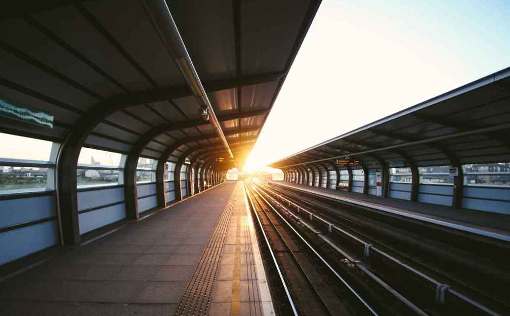 De zon schijnt over een leeg treinstation. Tussen twee overkapte perrons lopen de sporen ver naar voren.